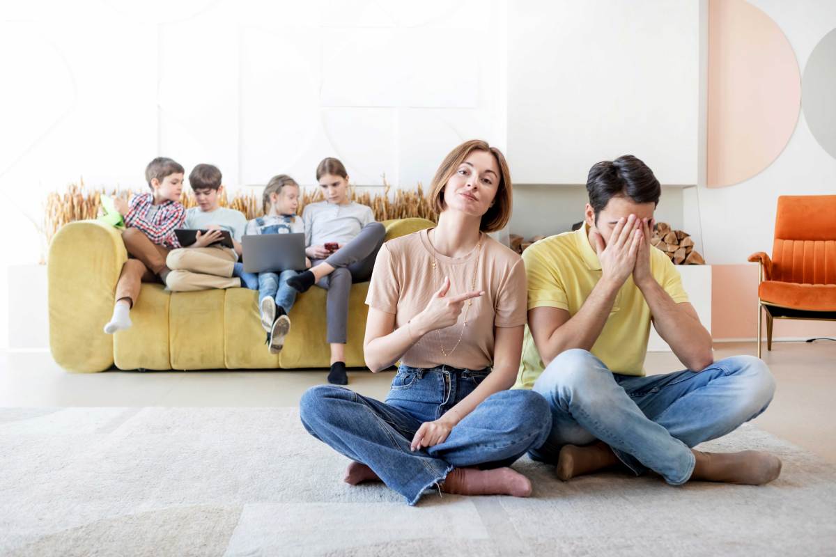 Frau und Mann sitzen auf dem Boden, im Hintergrund sieht man vier Kinder auf einem Sofa sitzen.