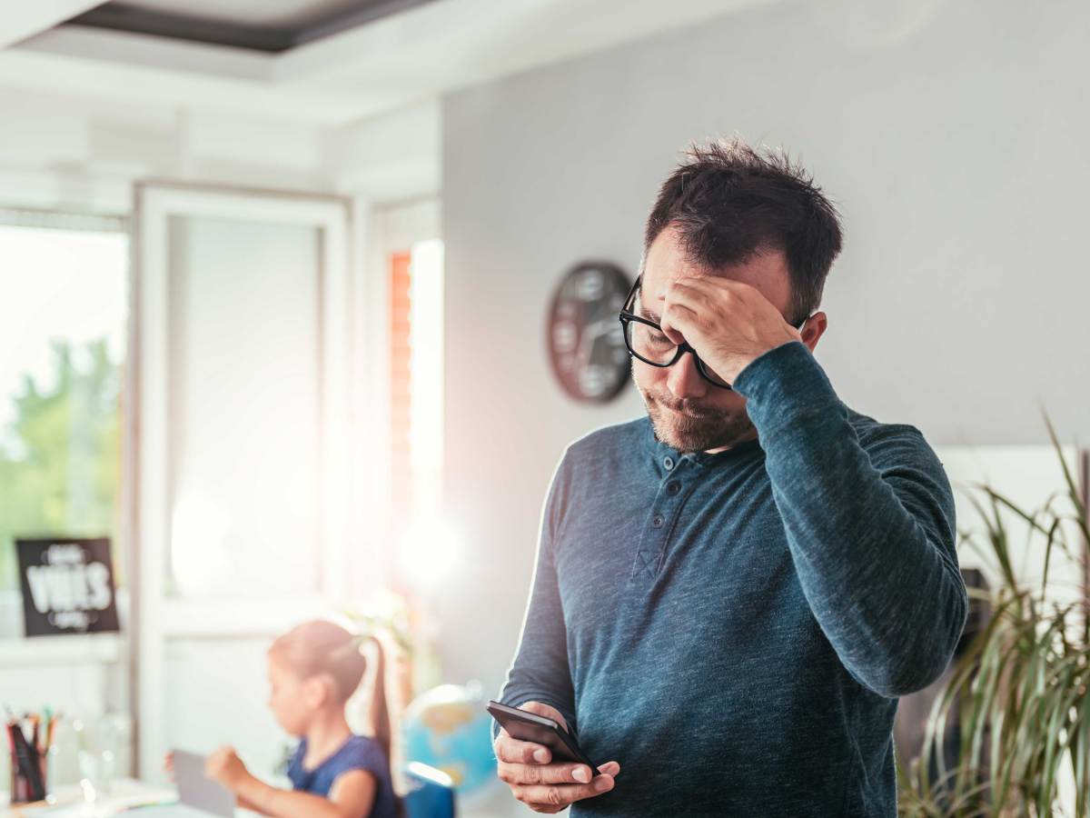 Vater schaut betroffen auf sein Smartphone, während die Tochter im Hintergrund am Tablet arbeitet.
