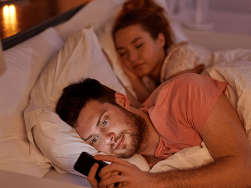 Mann, der im Bett an seinem Handy spielt, während im Hintergrund eine Frau im Bett schläft
