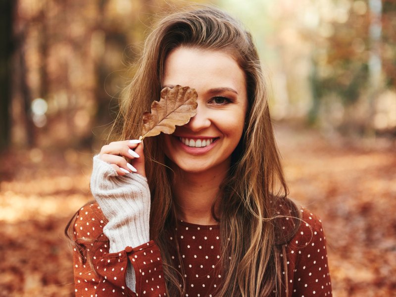 Brünette Frau, die dem Herbsttyp entspricht, steht in herbstlichem Wald und hält ein Laubblatt vor ihr Auge