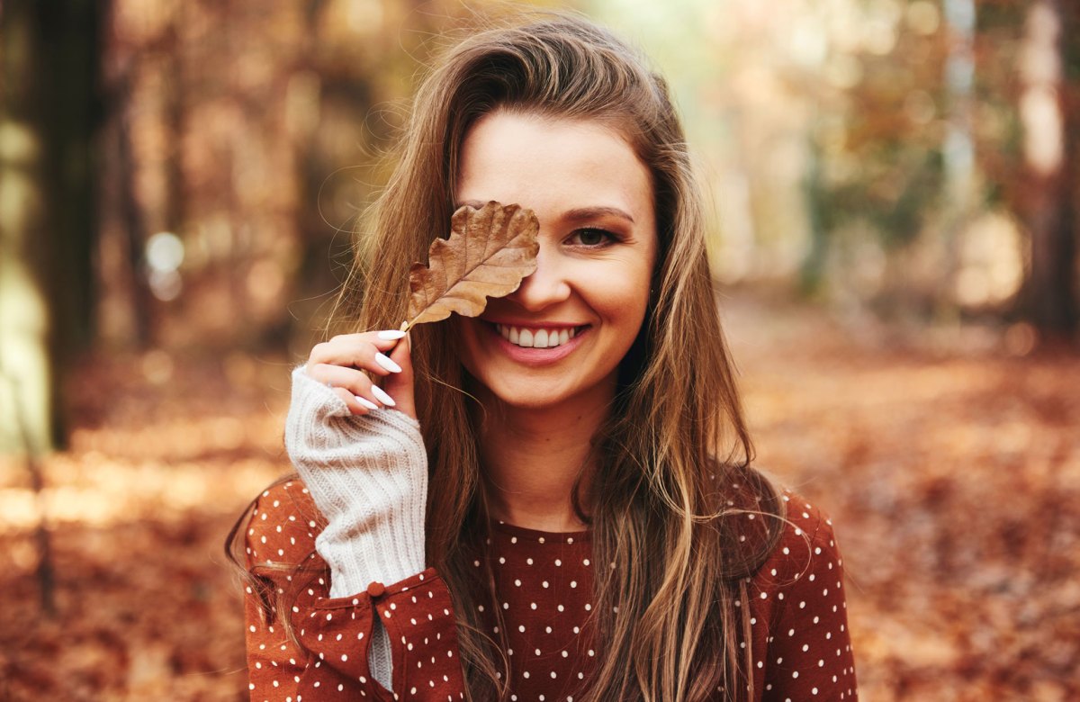 Brünette Frau, die dem Herbsttyp entspricht, steht in herbstlichem Wald und hält ein Laubblatt vor ihr Auge