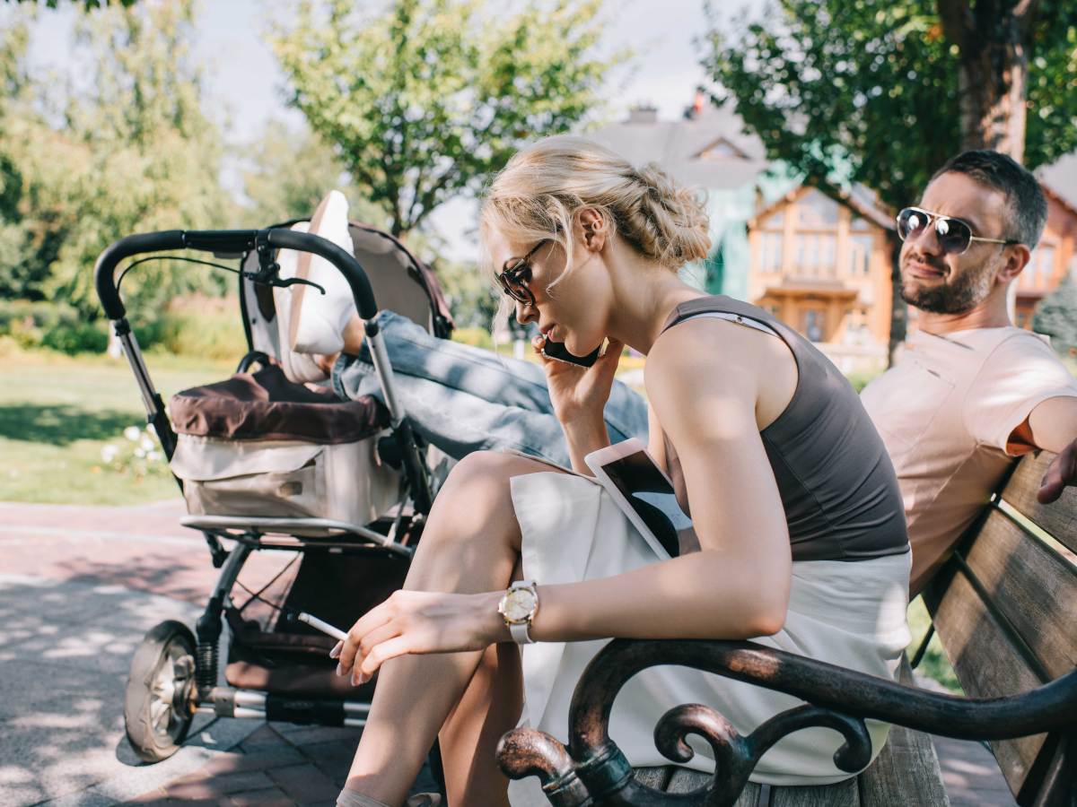 Paar sitzt auf einer Parkbank, er hat die Beine lässig auf dem Kinderwagen abgelegt, sie sitzt neben ihm, raucht und telefoniert am Handy.