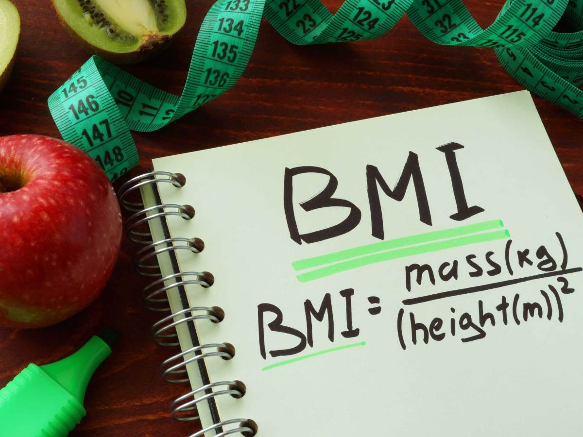 Notizbuch mit "BMI" und Formel, umgeben von Apfel, Kiwi und Maßband auf Holztisch.