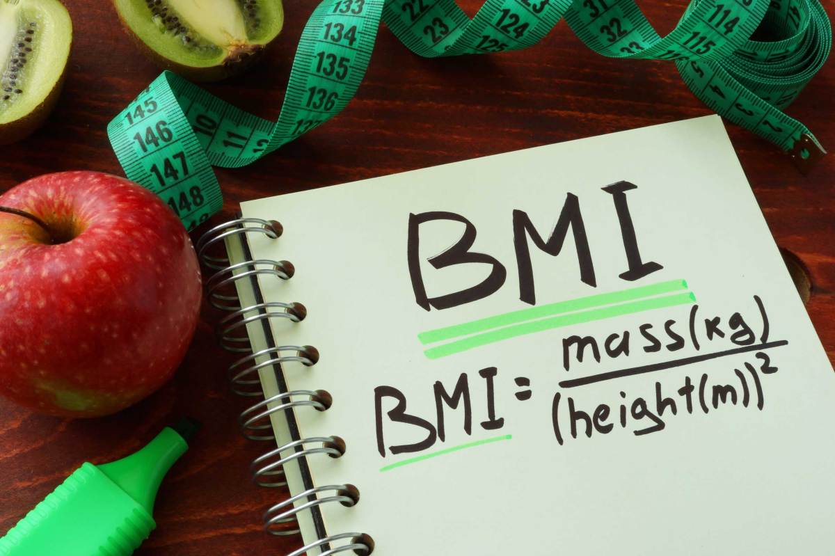 Notizbuch mit "BMI" und Formel, umgeben von Apfel, Kiwi und Maßband auf Holztisch.