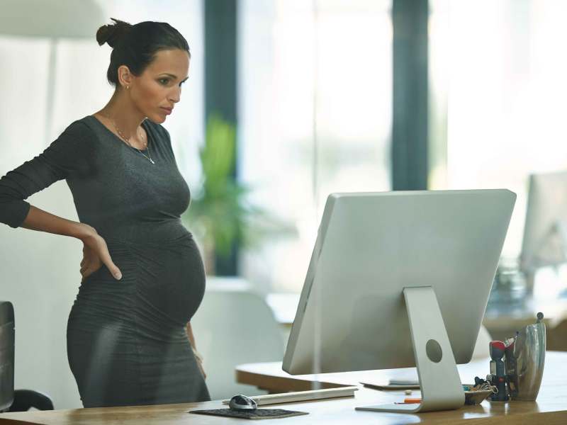 Schwangere Frau steht im Büro und schaut auf ihren PC-Bildschirm.