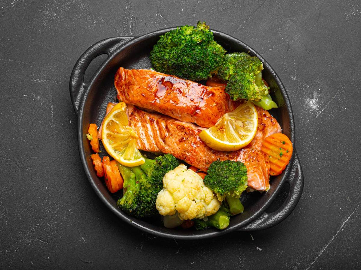 Gesunder gebackener Fisch Lachssteaks, Brokkoli, Blumenkohl, Karotten in Gusseisen Auflaufform Schüssel schwarzen dunklen Stein Hintergrund.