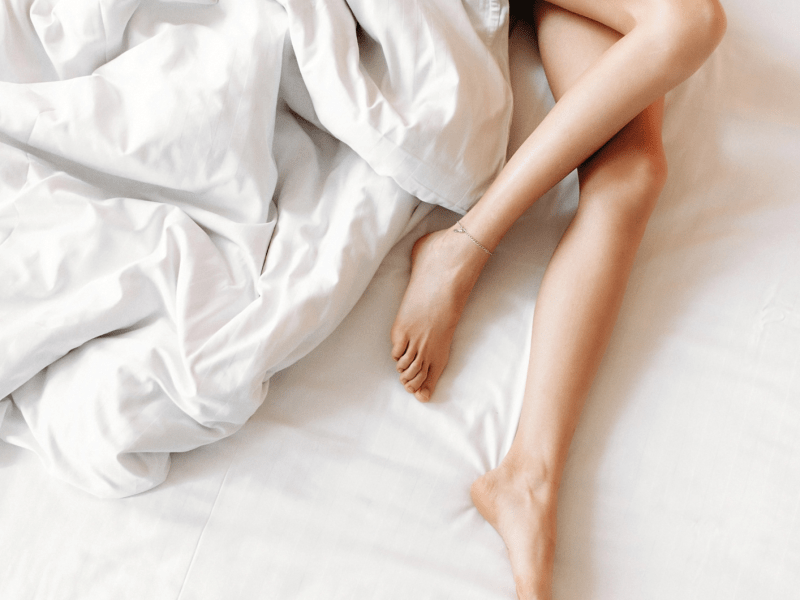 Frauenbeine im Bett auf einem weißen Laken, umhüllt von einer weißen Bettdecke