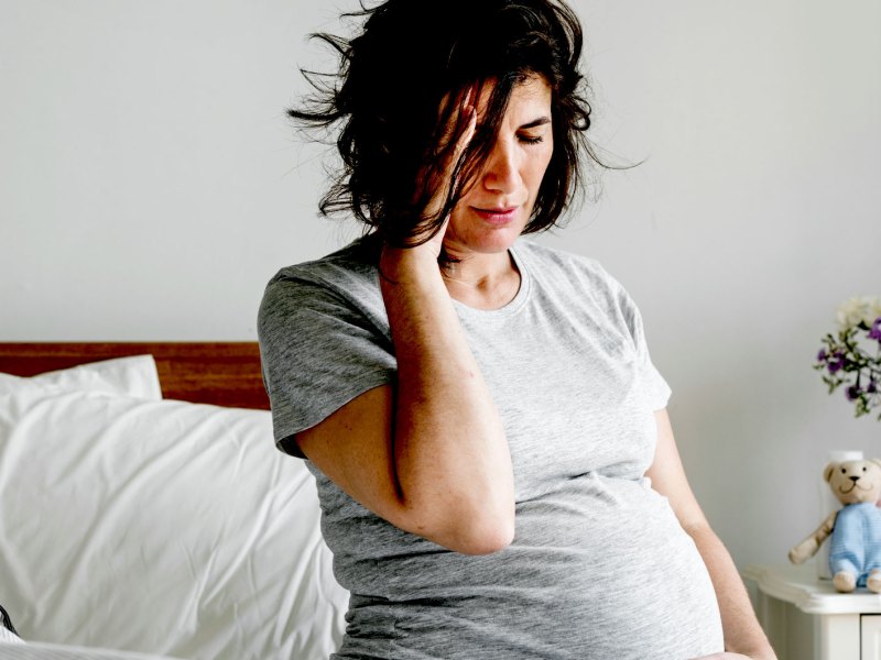 Schwangere Frau sitzt auf dem Bett, hält ihren Bauch mit der einen und ihren Kopf mit der anderen Hand fest. Das Gesicht ist leicht schmerzverzerrt.