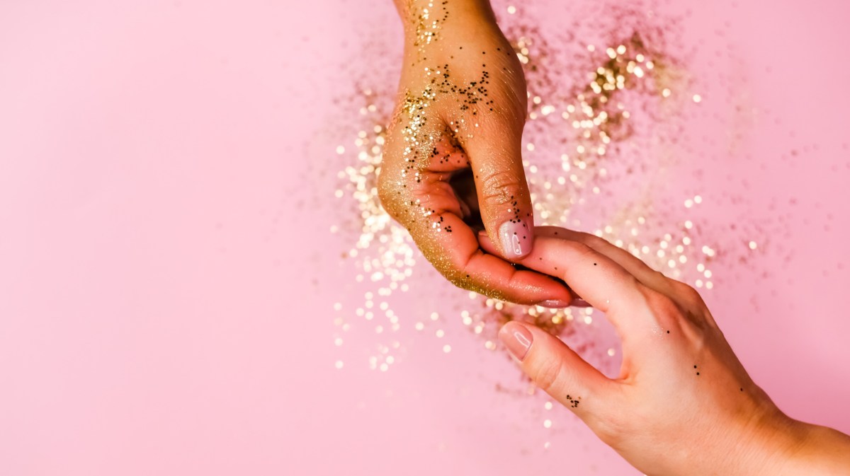 Zwei Hände mit GLitzer vor rosa Hintergrund, die sich an den Fingerspitzen berühren und anfassen