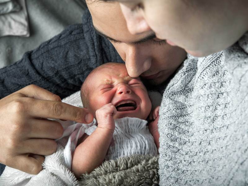 Nahaufnahme eines weinenden Neugeborenen. Die Eltern sind angeschnitten zu erkennen.
