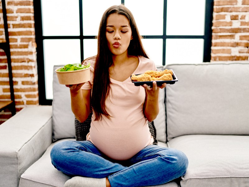 Schwangere Frau sitzt im Schneidersitz auf dem Sofa, hält zwei Teller in der Hand und schaut unentschlossen zwischen einem Salat und frittiertem Fisch hin und her.