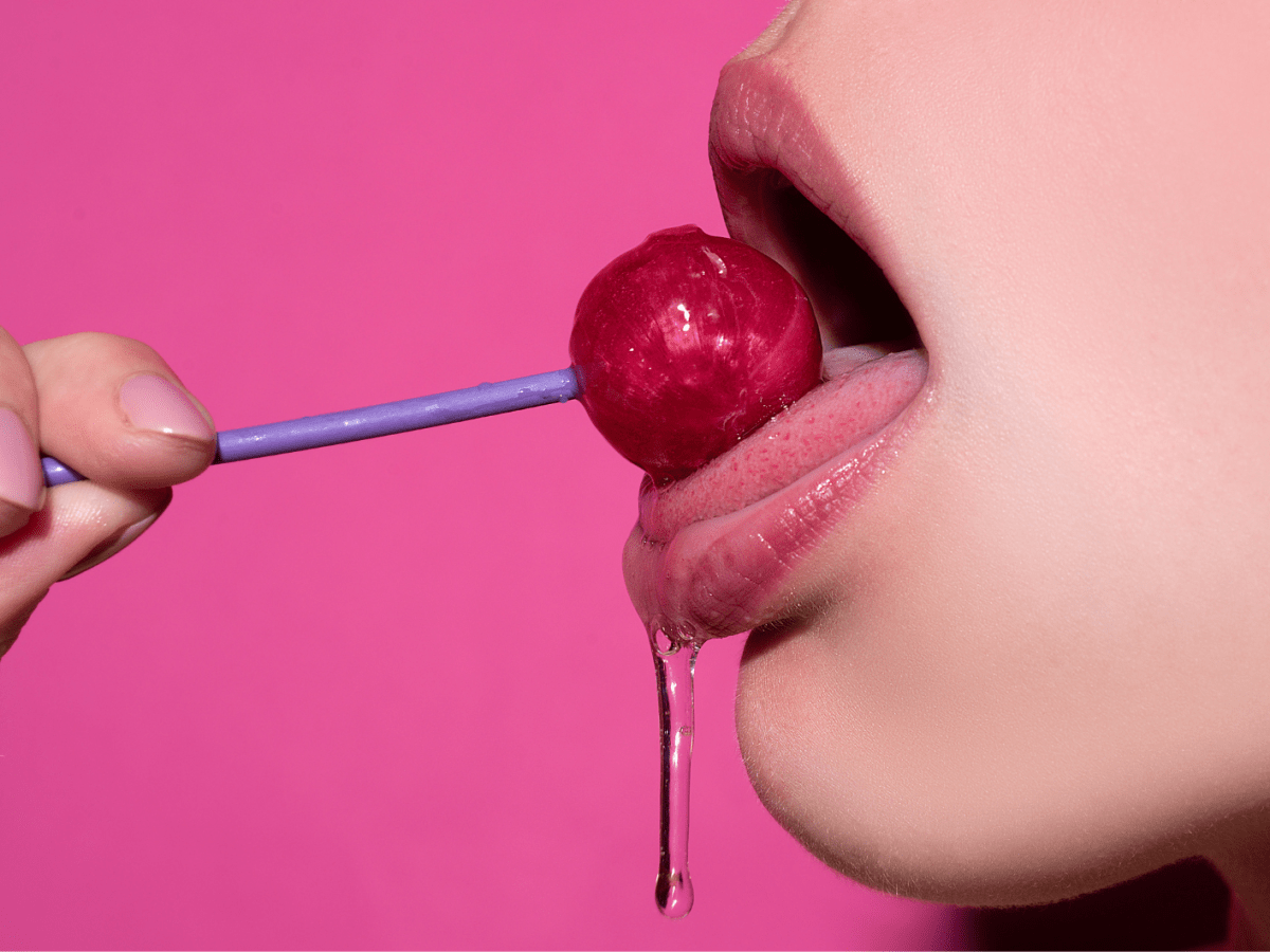 Nahaufnahme eines geöffneten Mundes mit glänzenden Lippen, der einen roten Lolli am lilafarbenen Stiel leckt, vor pinkem Hintergrund.