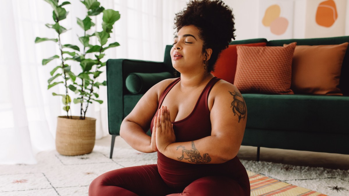 Frau vor Couch, die Yoga macht und die Hände vor der Brust zusammenlegt