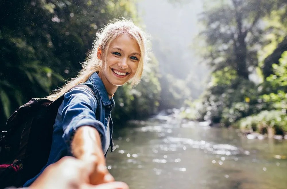 Frau an einem Fluss, die lächelnd in die Kamera blickt und ihre Hand Richtung Kamera ausstreckt