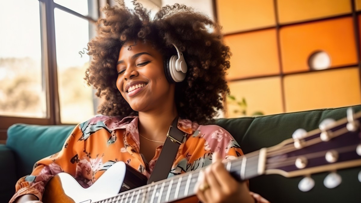 Afro-amerikanische Frau mit Gitarre in der Hand und Kopfhörern auf, die glücklich und entspannt auf einem Sofa sitzt
