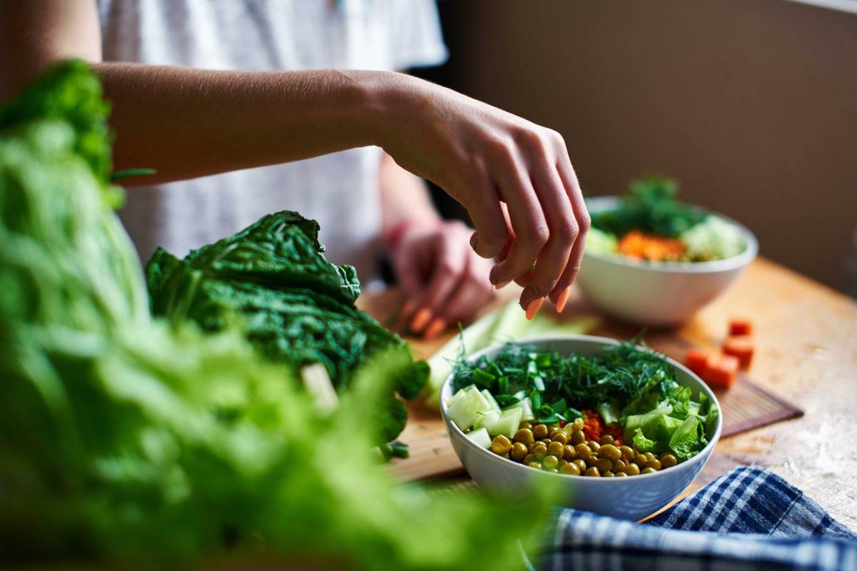 Weibliche Hand gibt grüne Zwiebeln in eine Schüssel mit grünen Erbsen, Gurken, Karotten, Salat und Dill, die auf einem Tisch stehen