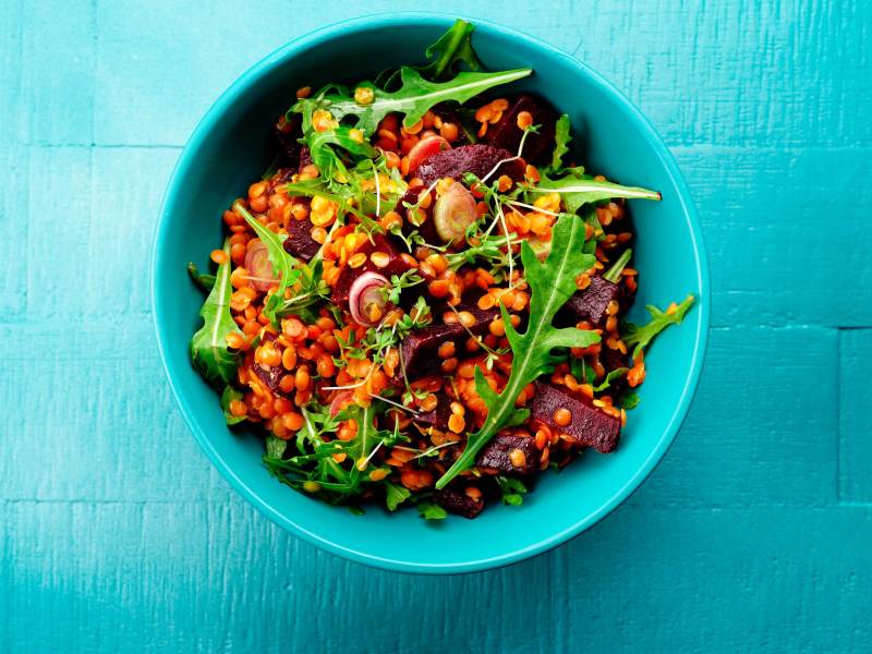 Türkise Schale mit gesundem Salat aus Linsen, Rote Beete und Rucola, angerichtet auf blauem Holztisch.