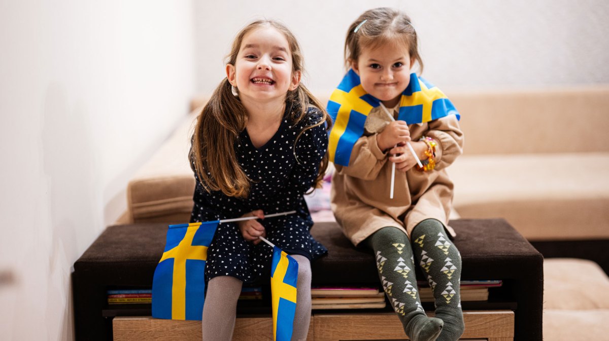 Zwei kleine Mädchen sitzen auf einer großen Kiste und halten je zwei schwedische Flaggen in ihren Händen.