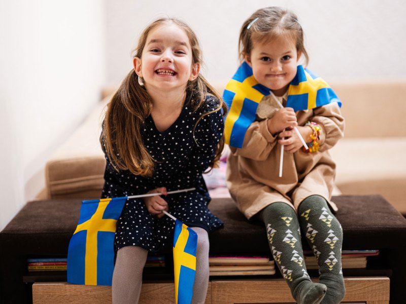 Zwei kleine Mädchen sitzen auf einer großen Kiste und halten je zwei schwedische Flaggen in ihren Händen.