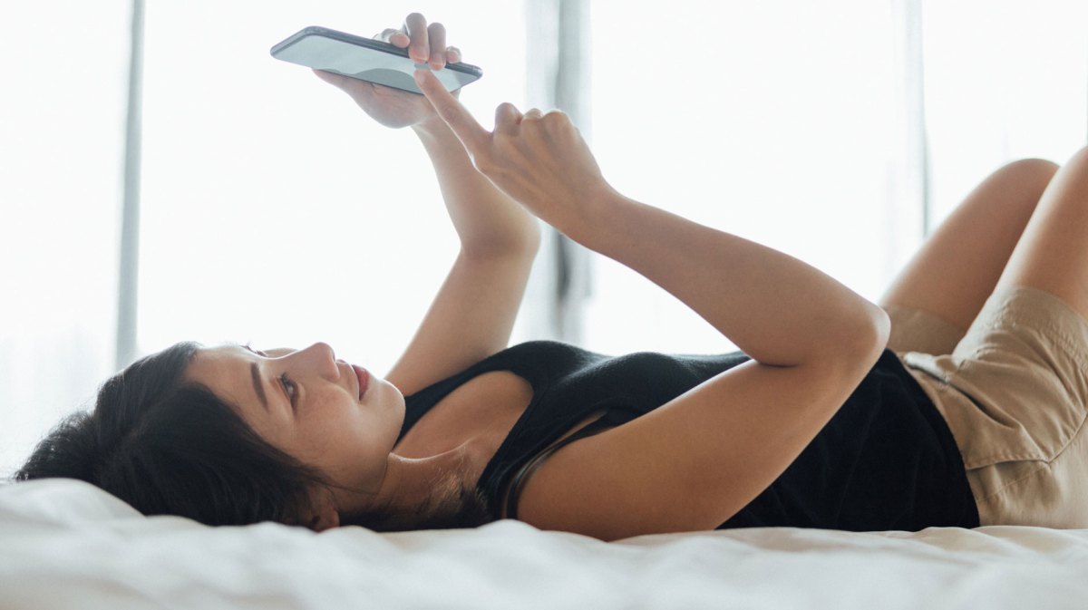 Frau liegt mit einem Smartphone in der Hand auf dem Bett