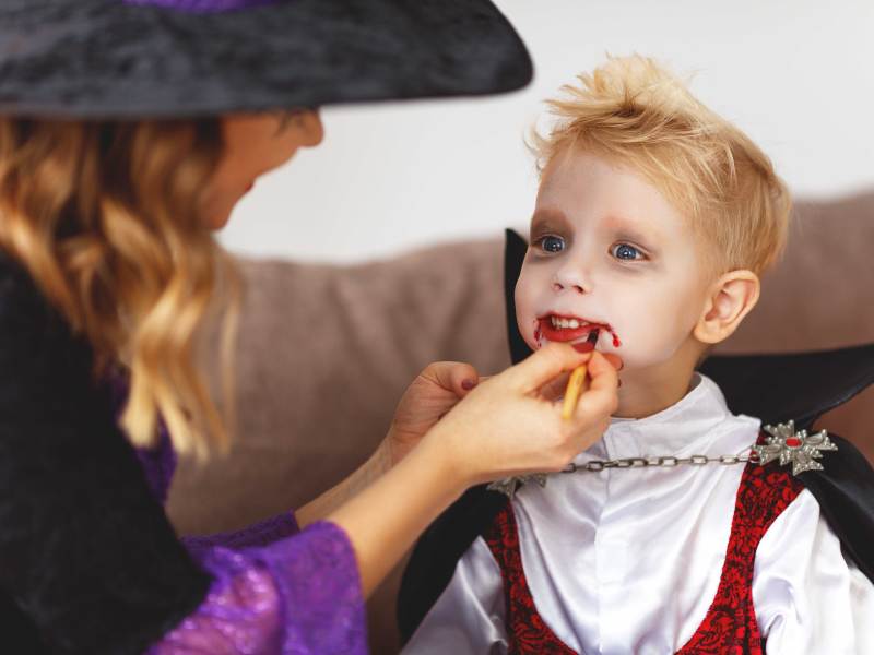 Junge wird von seiner Mutter, die ein Hexenkostüm trägt, zu einem Vampir geschminkt.