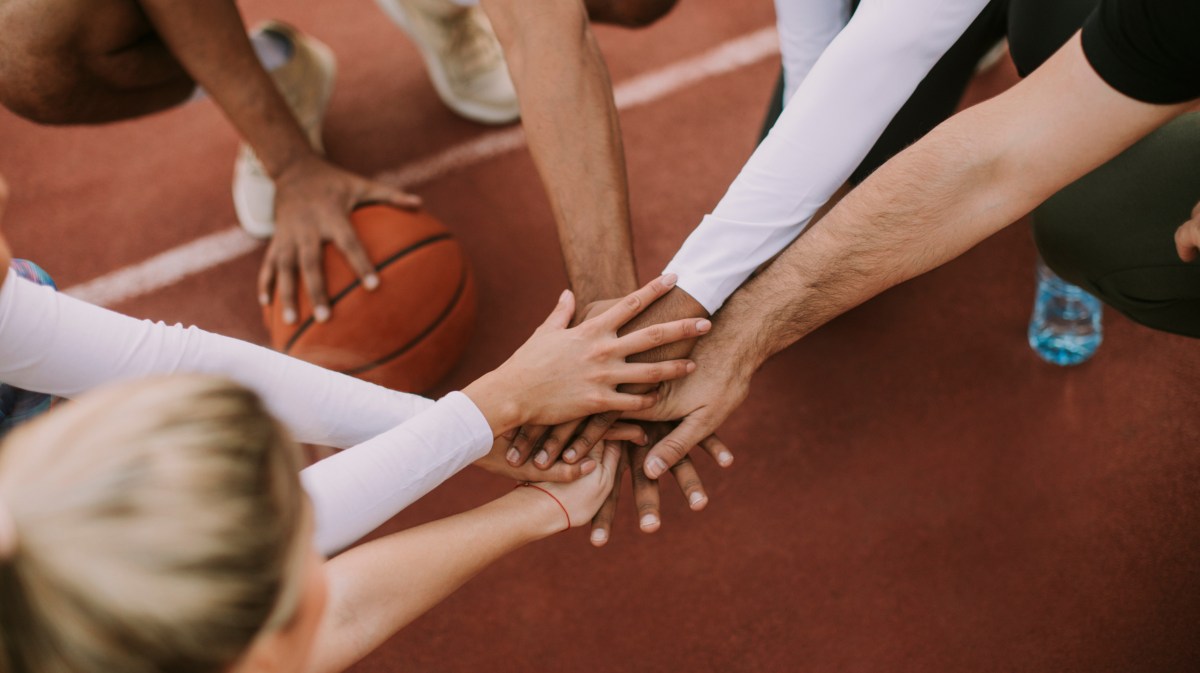 Menschen hocken auf einem Basketballplatz im Kreis und legen in der Mitte die Hände zusammen.