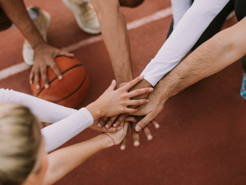 Menschen hocken auf einem Basketballplatz im Kreis und legen in der Mitte die Hände zusammen.