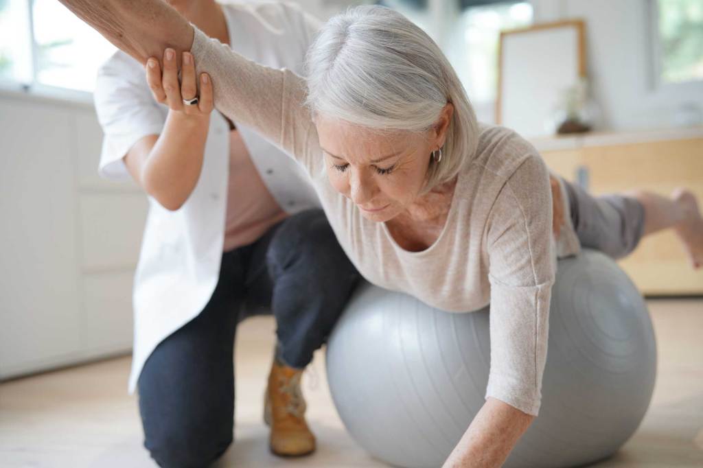 Ältere Frau mit grauem Haar macht mithilfe einer Physiotherapeutin Übungen auf einem Gymnastikball.