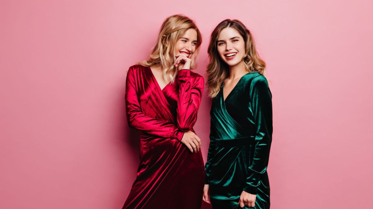 Zwei junge Frauen stehen vor rosanen Hintergrund, eine trägt ein rotes Kleid, die andere ein grünes.