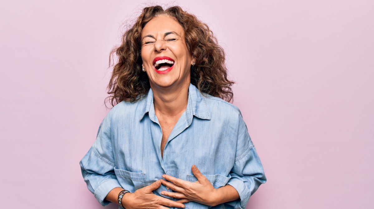Frau mittleren Alters vor einem rosa Hintergrund lacht mit offenem Mund und hält die Hände vor den Bauch vor Lachen.