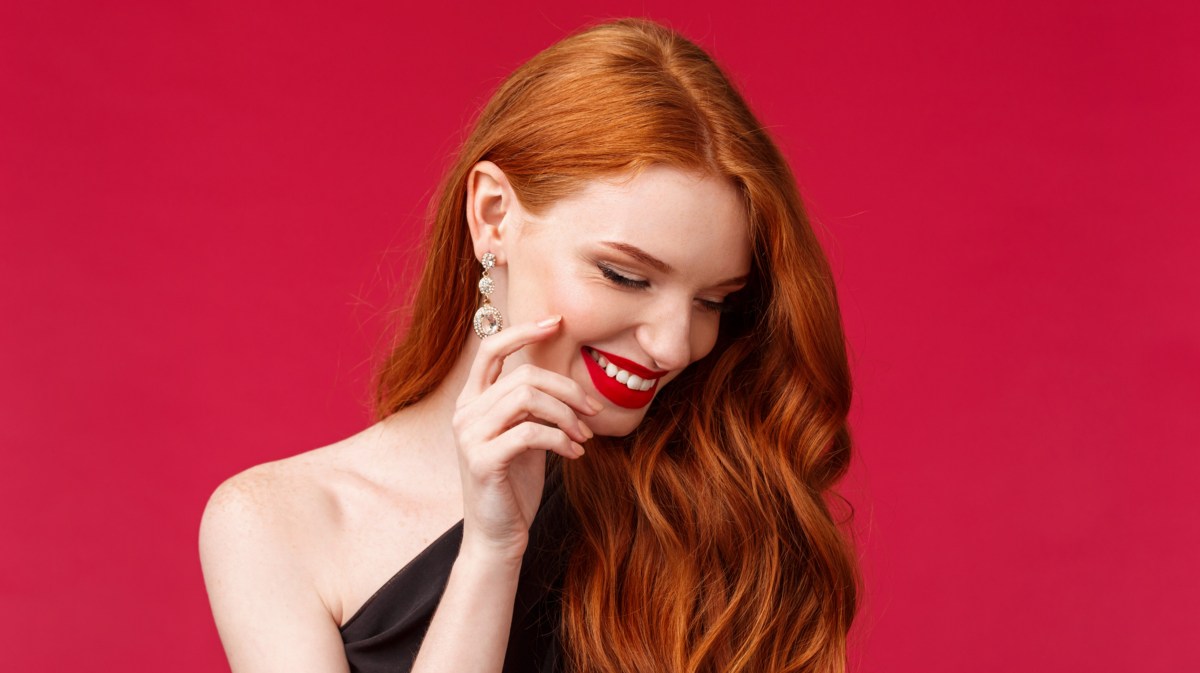Frau mit roten Haaren vor rotem Hintergrund, die zur Seite lächelt und ihre Hand schüchtern an den Mund hält