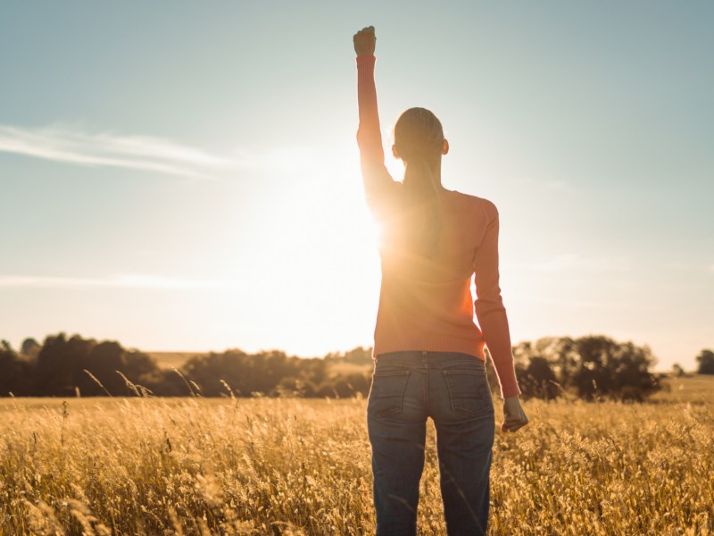 Frau auf einem Feld im Sonnenaufgang, die ihre rechte Hand in die Luft hält