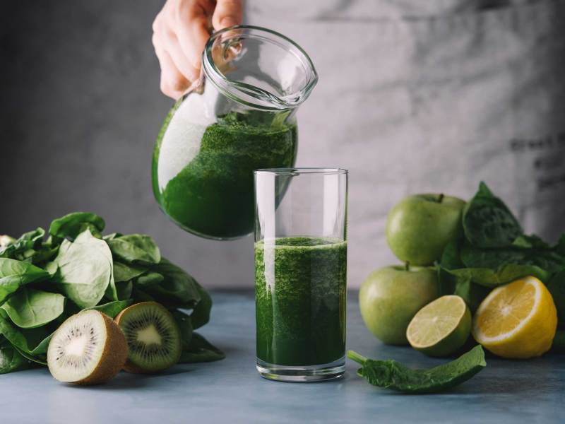 Weibliche Hände gießen grünen Smoothie in ein Glas. Zutaten: Spinat, Apfel, Kiwi, Zitrone, Limette. Detox-Frühstück