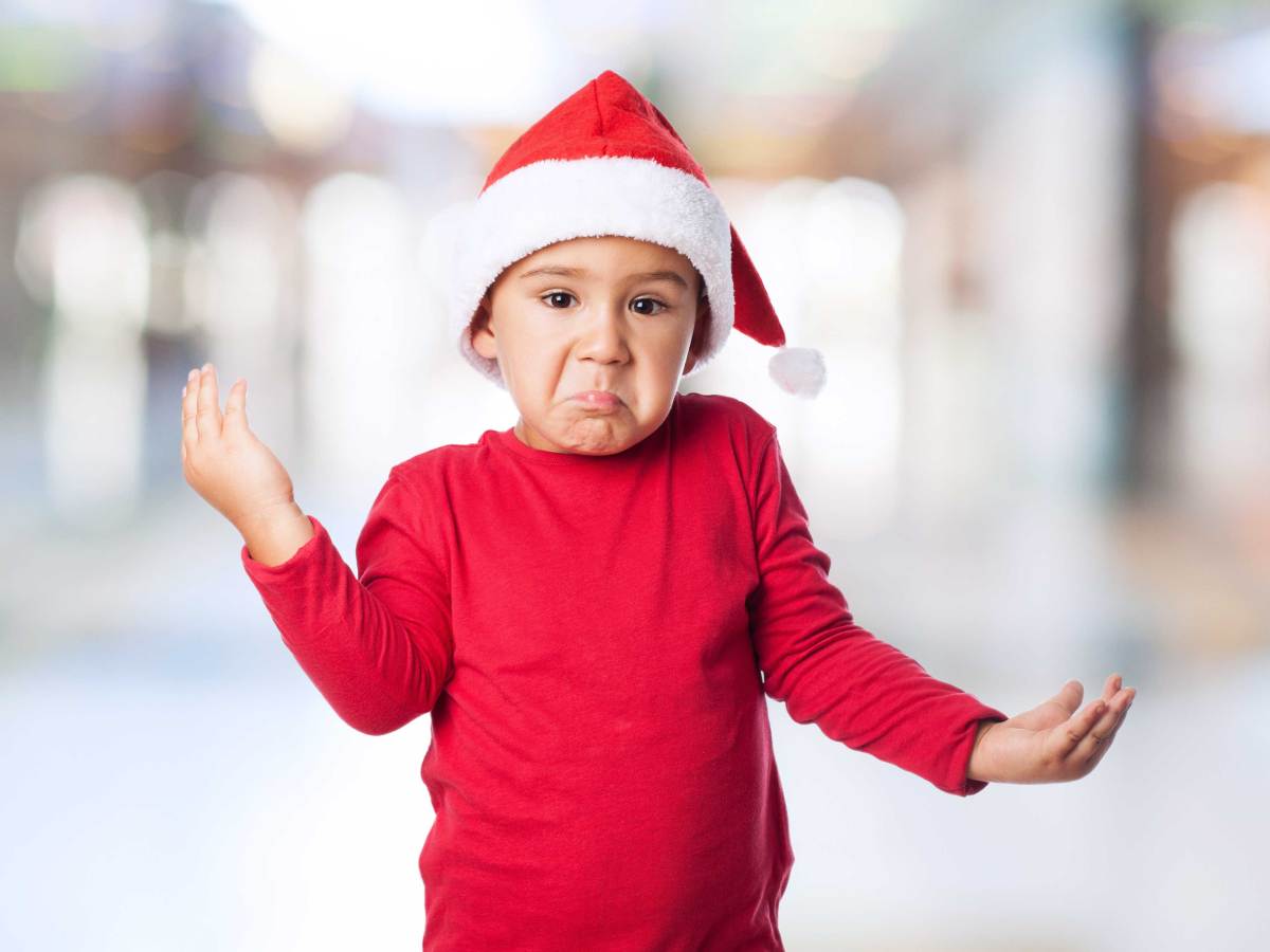 Kleiner Junge in rotem Pullover und mit Weihnachtsmannmütze schaut unsicher in die Kamera und zuckt mit den Schultern.