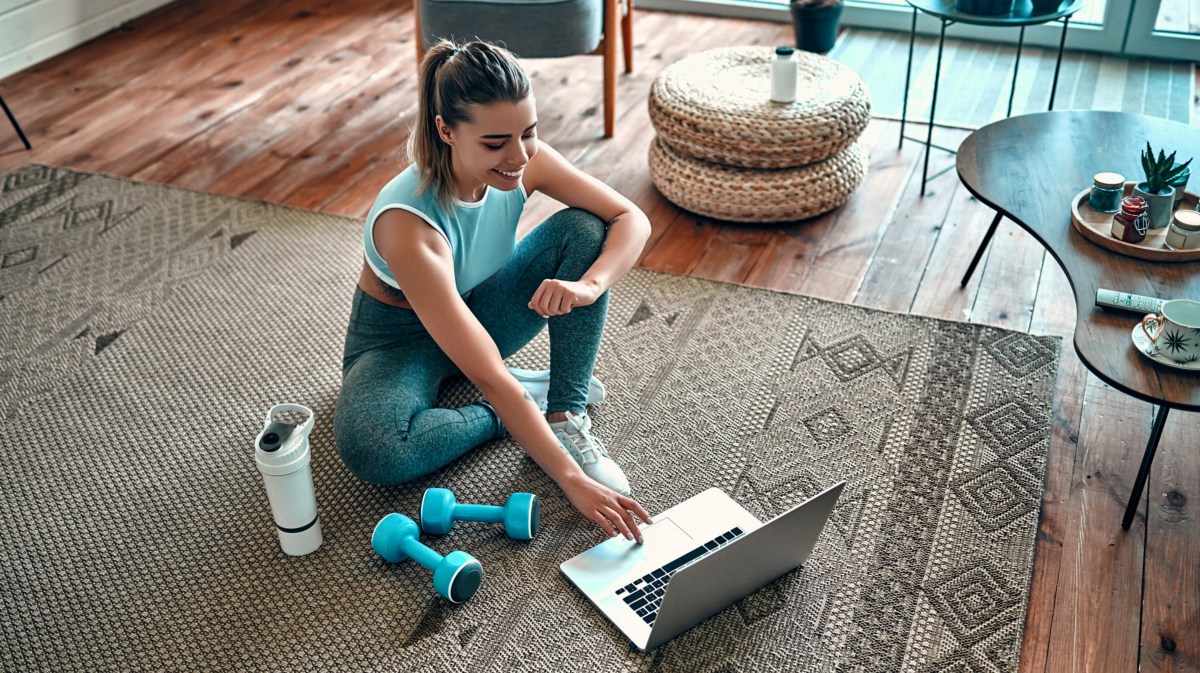 Frau sitzt mit Fitness-Geräten auf dem Boden vor ihrem Laptop