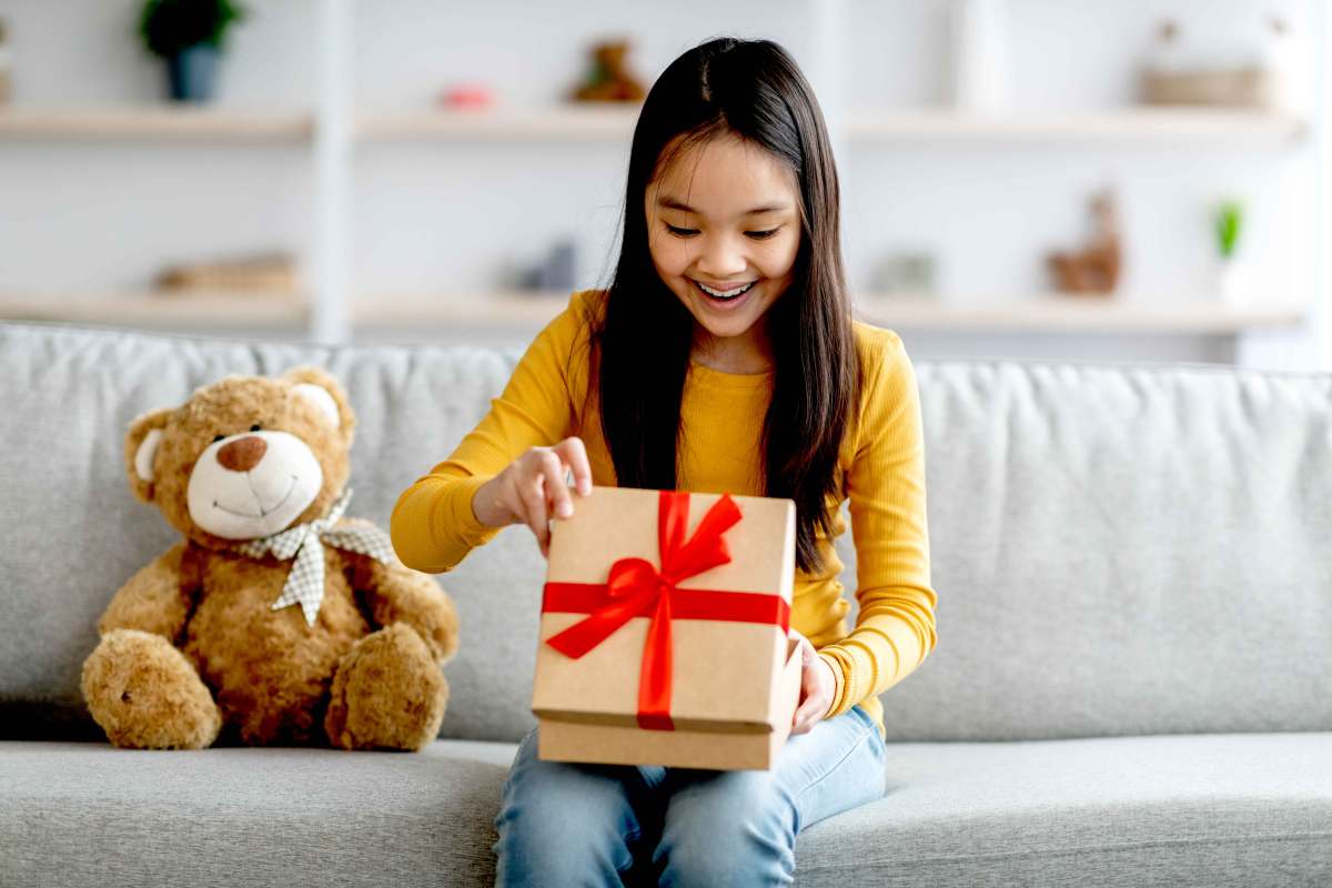 Mädchen sitzt auf der Couch und schaut freudig überrascht in eine Geschenkbox.