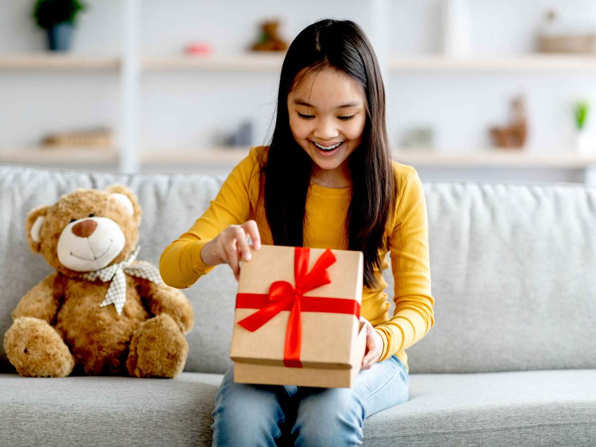 Mädchen sitzt auf der Couch und schaut freudig überrascht in eine Geschenkbox.