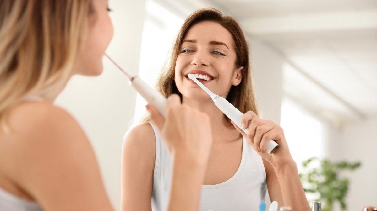 Frau putzt sich vor Spiegel mit elektrischer Zahnbürste die Zähne.