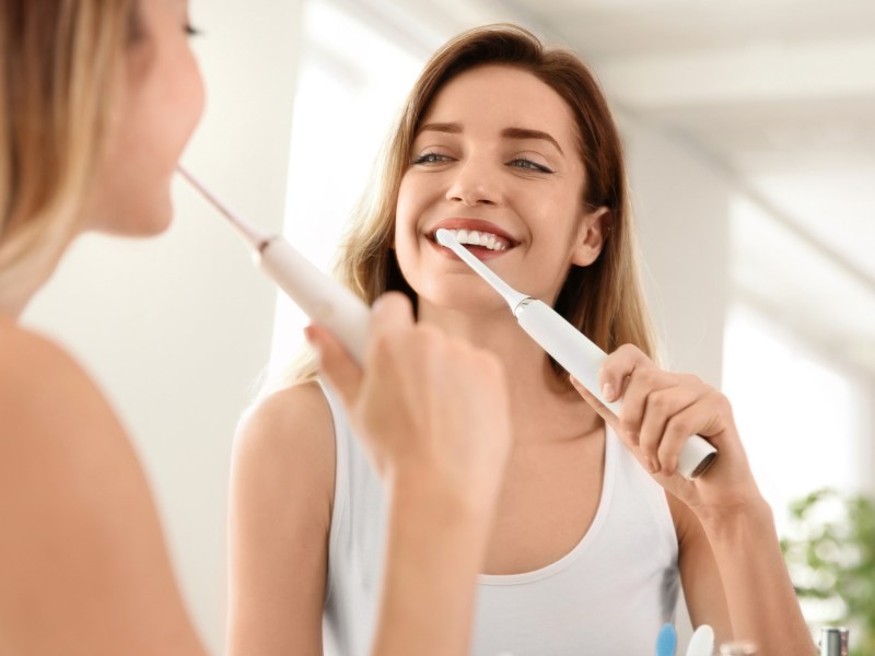 Frau putzt sich vor Spiegel mit elektrischer Zahnbürste die Zähne.