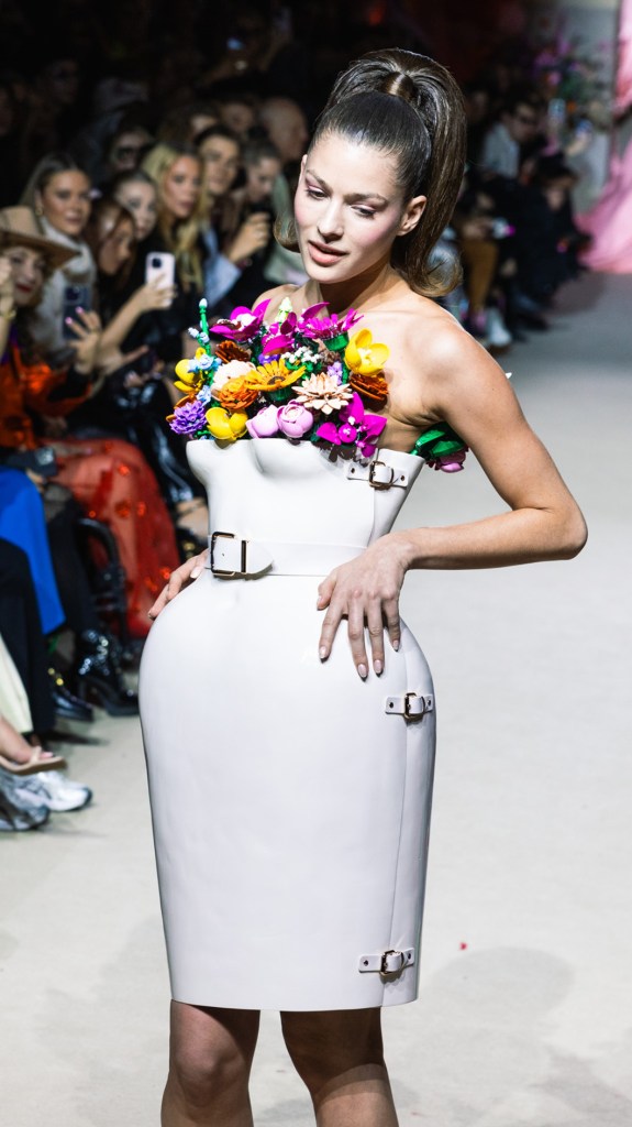 Eine junge Frau mit hohem Pferdeschwanz läuft über den Laufsteg in einem weißen Korsettkleid mit bunten Lego Botanicals Blumen und Metallschnallen.