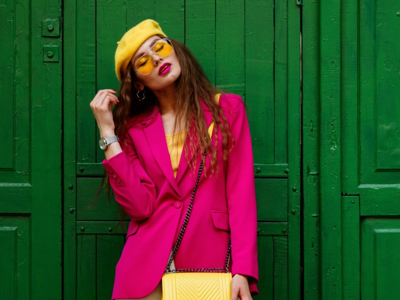Frau vor grünem Hauseingang in pinker Kleidung, mit Sonnenbrille und gelber Tasche