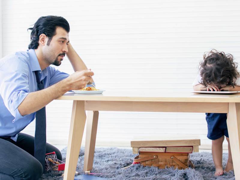 Vater schaut frustriert auf sein kleines Kind, das bockig den Kopf auf den Tisch gelegt hat, statt zu essen.