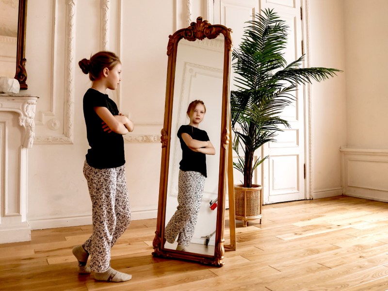 Junges Mädchen steht vor einem Ganzkörperspiegel und schaut mit verschränkten Armen hinein.