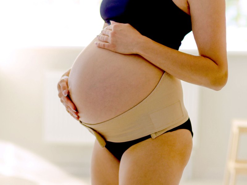 Hochschwangere Frau hat sich einen breiten Klettgürtel um das Becken gelegt, welcher den großen Babybauch stützt.