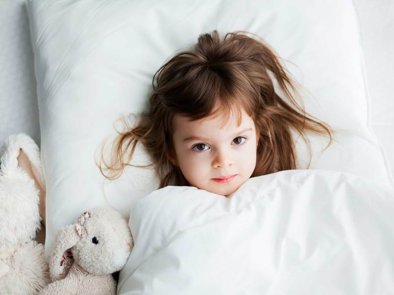 Zirka vierjähriges Mädchen liegt zugedeckt und hellwach im Bett.