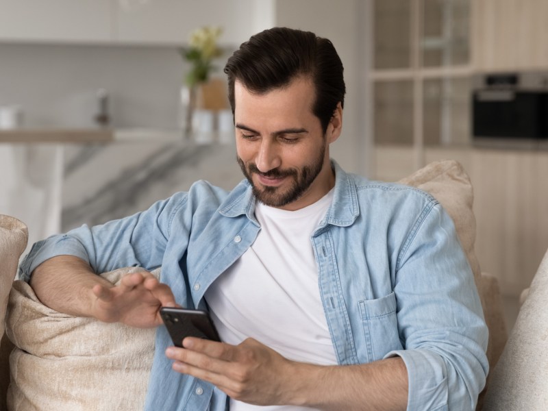 Mann der auf der Couch sitzt und sein Handy in der Hand hält, während er lächelt