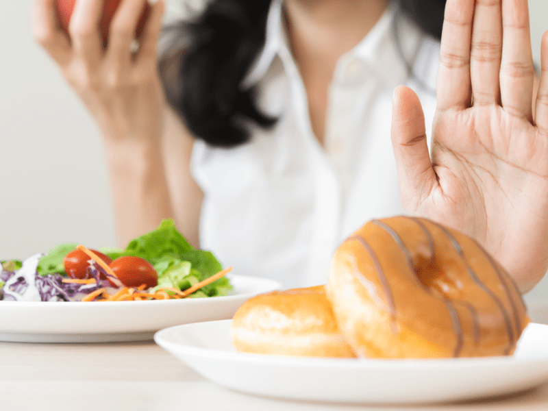 Frauenhand, die einen Salatteller vor sich hat und einen Teller voller Donuts zur Seite schiebt