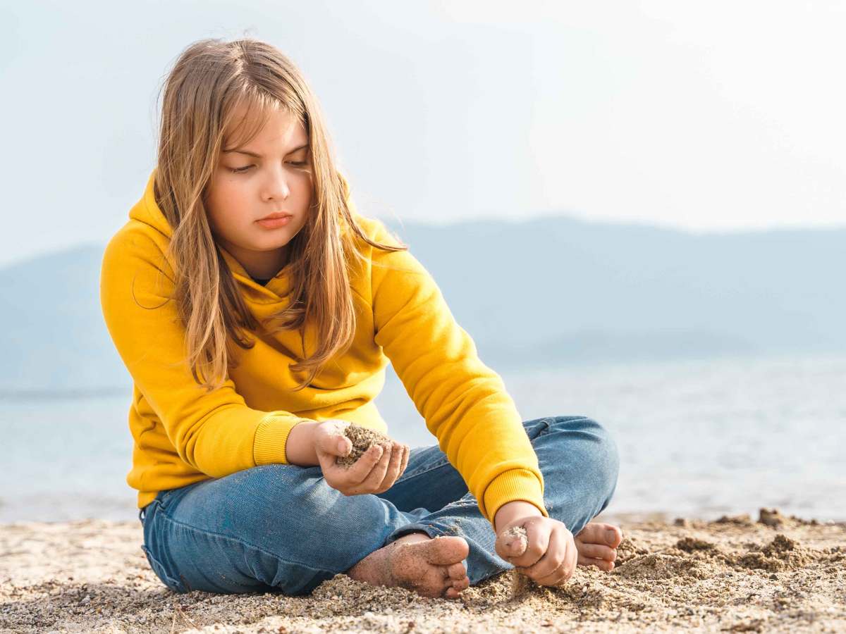 Mädchen um die 12 Jahre sitzt in Jeans und Hoodie alleine am Strand und spielt gedankenversunken mit dem Sand.