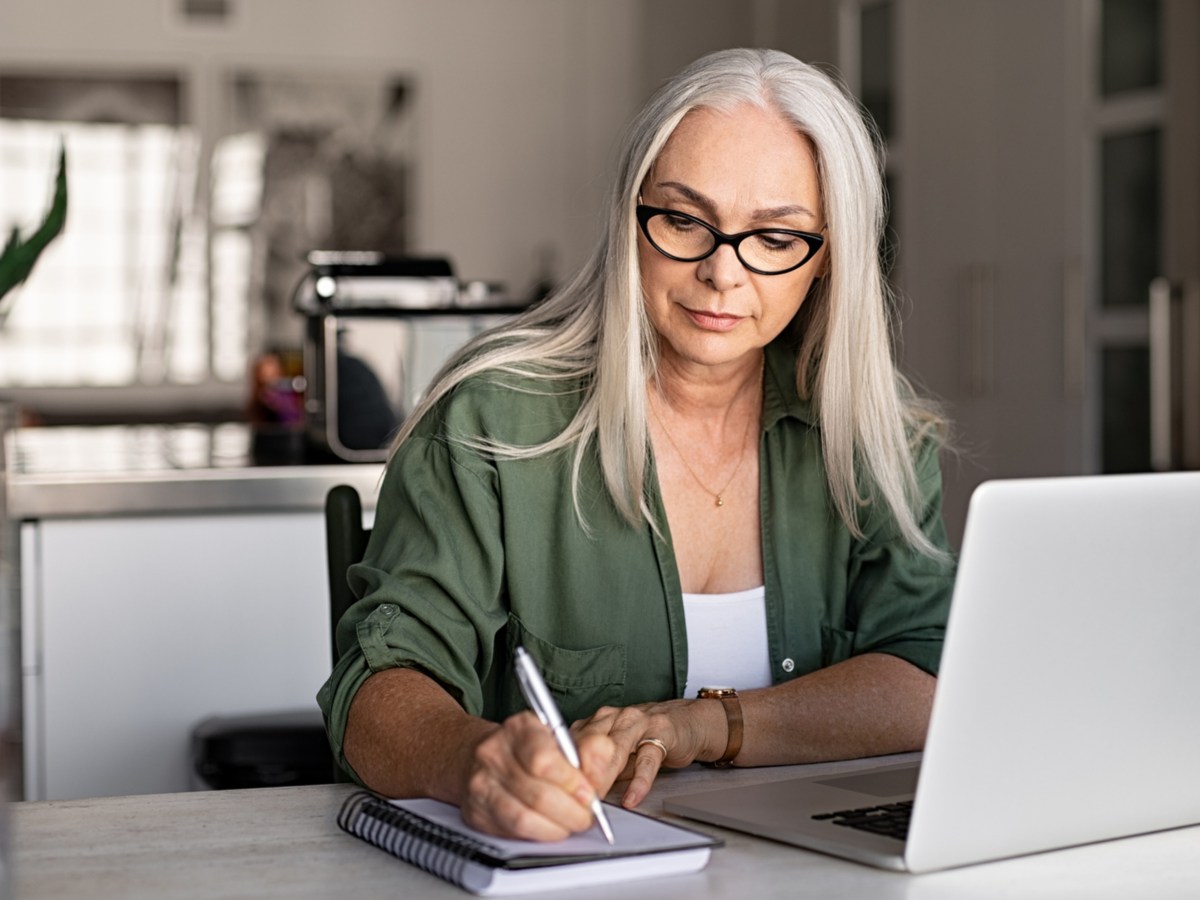 Frau mittleren Alters mit langen weißen Haaren schreibt etwas in ein Haushaltsbuch und sitzt vor ihrem Laptop.