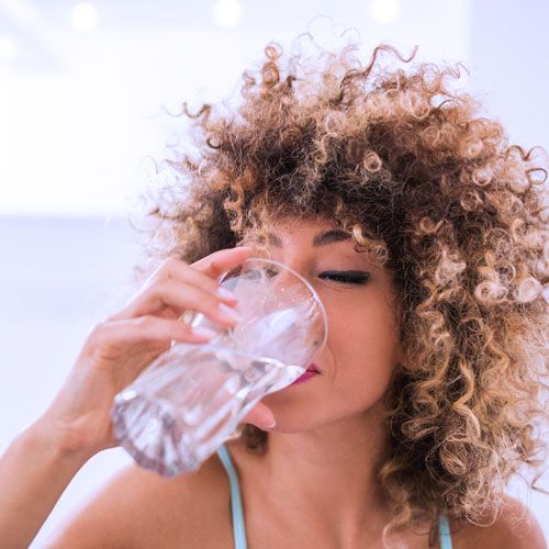 Tipps gegen einen Blähbauch: Viel stilles Wasser trinken Frau mit Locken trinkt aus einem Wasserglas